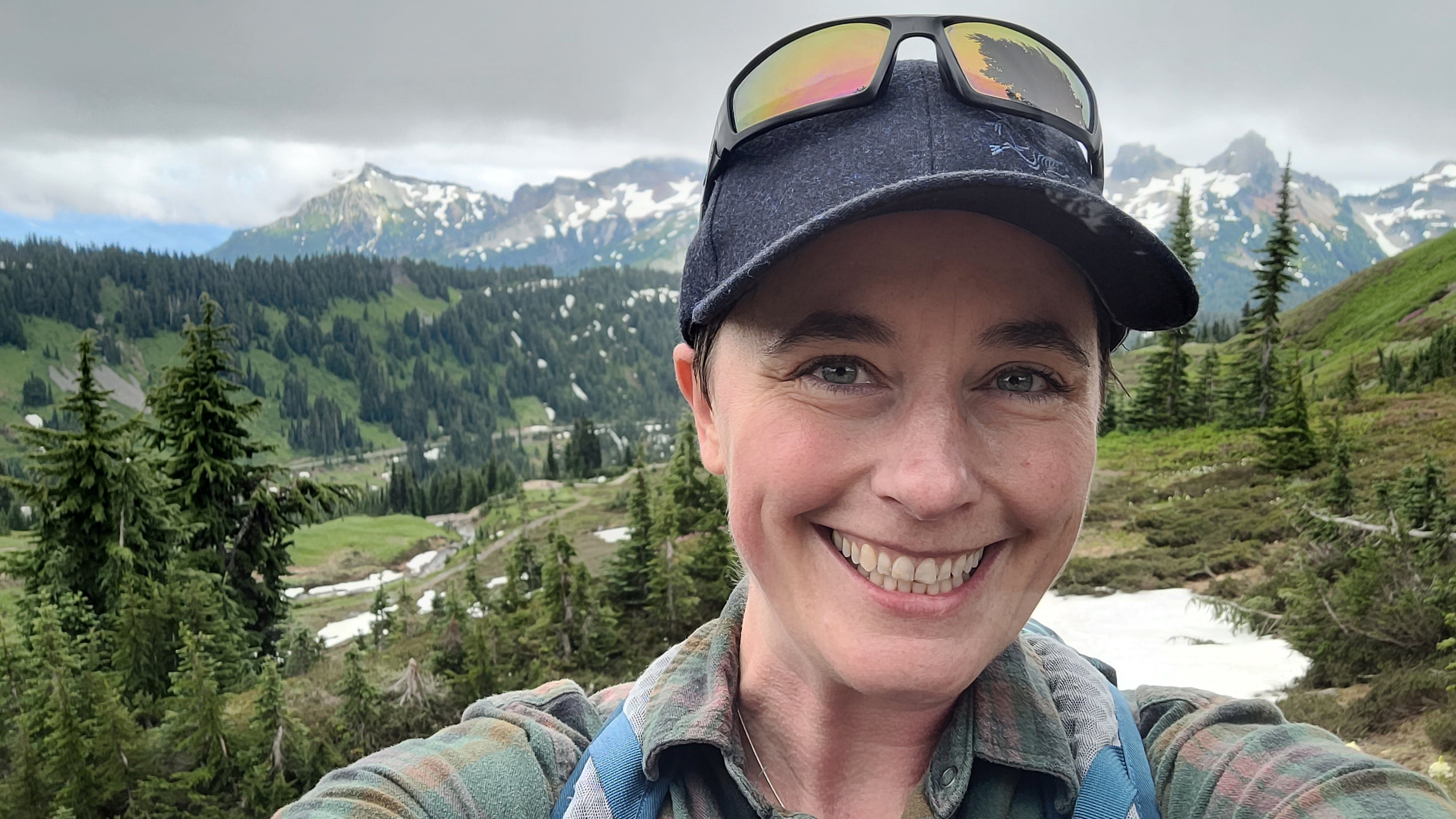 Smiling woman in charcoal grey baseball hat, amongst evergreen trees with Cascade mountains in the background.