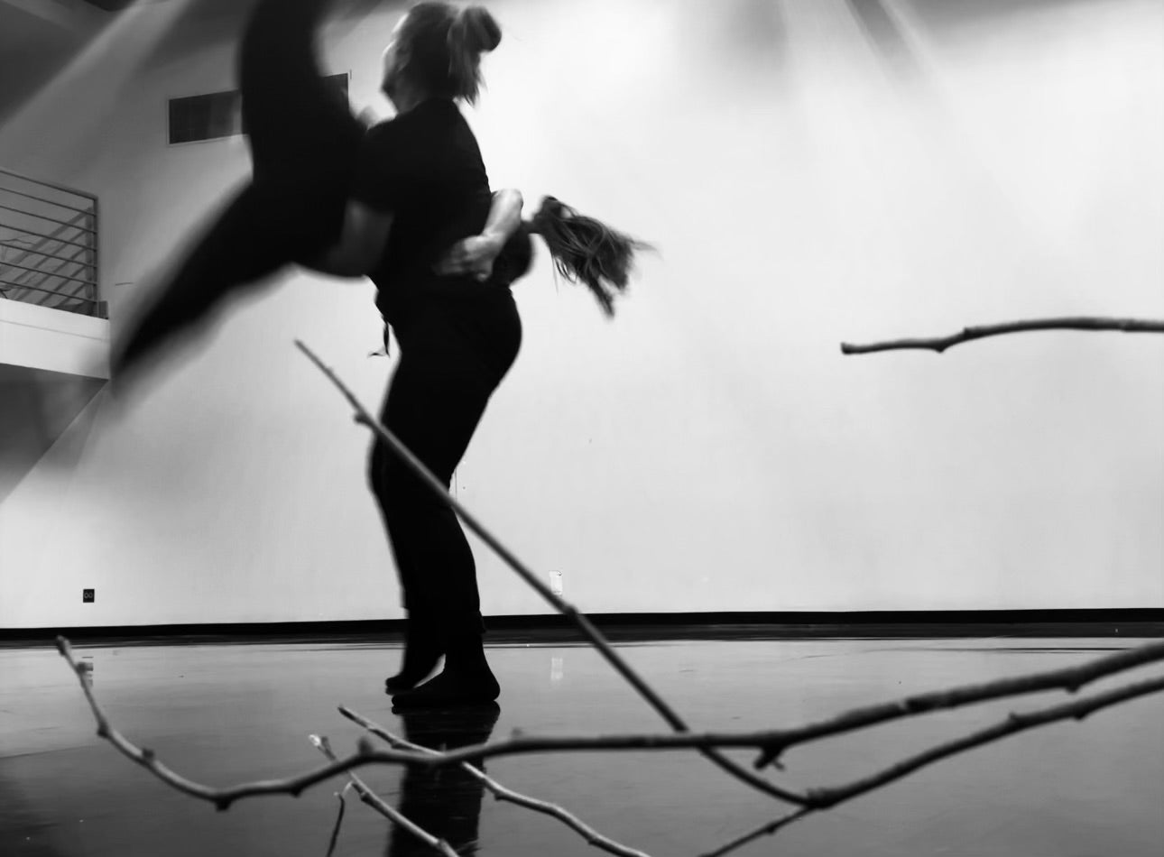 Nicole is supporting her dance partner in this black and white photography. Nicole stands facing the right side of the room as her partner, Zarina, swings her legs horizontally across Nicole's trunk. Zarina's legs split and are reaching past the frame of the photo. A barren tree branch lies on the floor in the foreground of the photograph taken by Haley Wilcox.