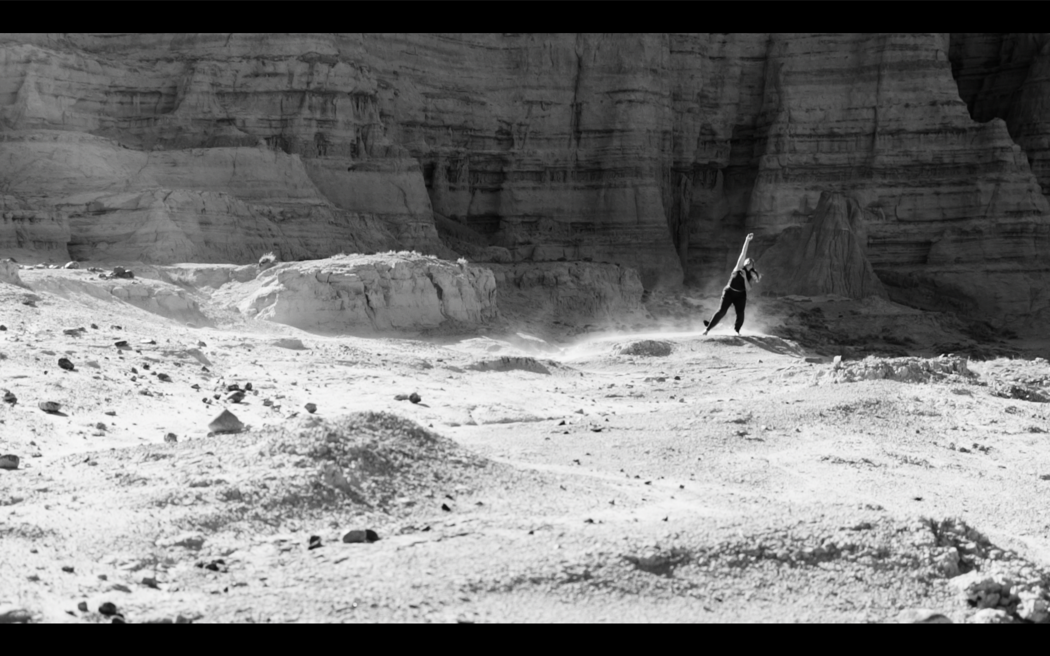 Nicole is standing among sandstone cliffs reaching her right arm upward and right leg to the side as the wind blows dust around her body. Black and white photo taken by Dmitri Von Klein.