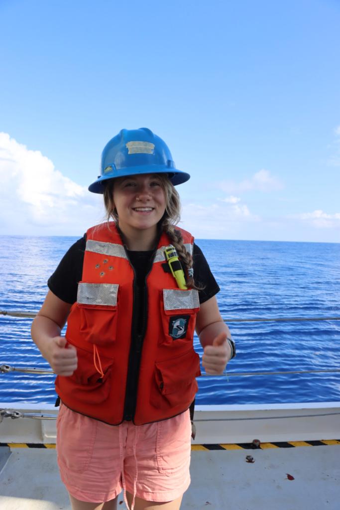 A researcher wearing a lifesaving vest and hard hat while sampling on the back of a ship. Smiling and with thumbs up.