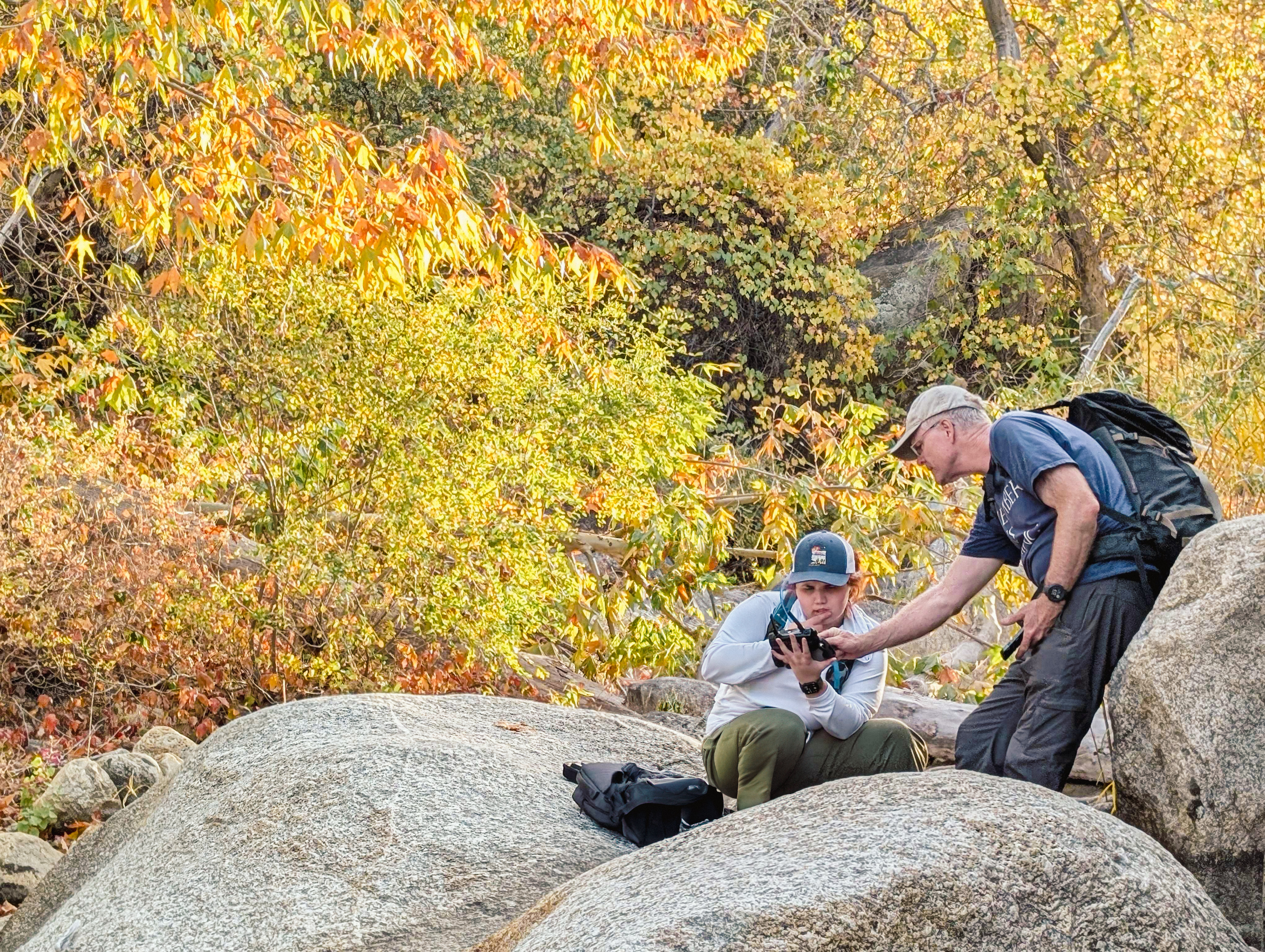 Emily looking at an ipad with Kelin Whipple (secondary advisor) pointing at something on the screen. They are sitting on large grey boulders with yellow and orange leaves from trees behind them. 