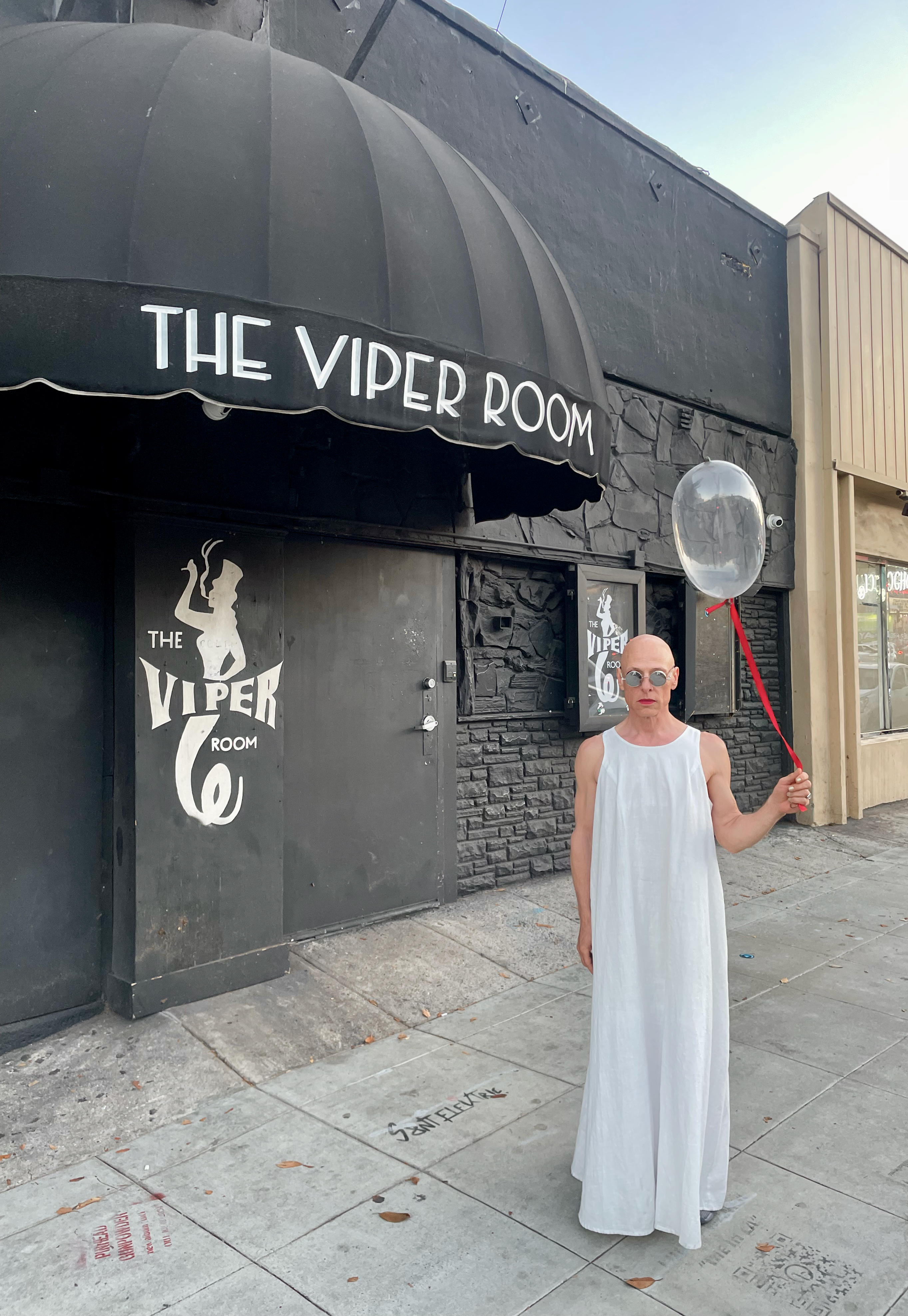a bald man in a white gown and holding a single floating condom attached with red ribbon while standing on the sidewalk in front of the viper room - a famed sunset strip nighclub.