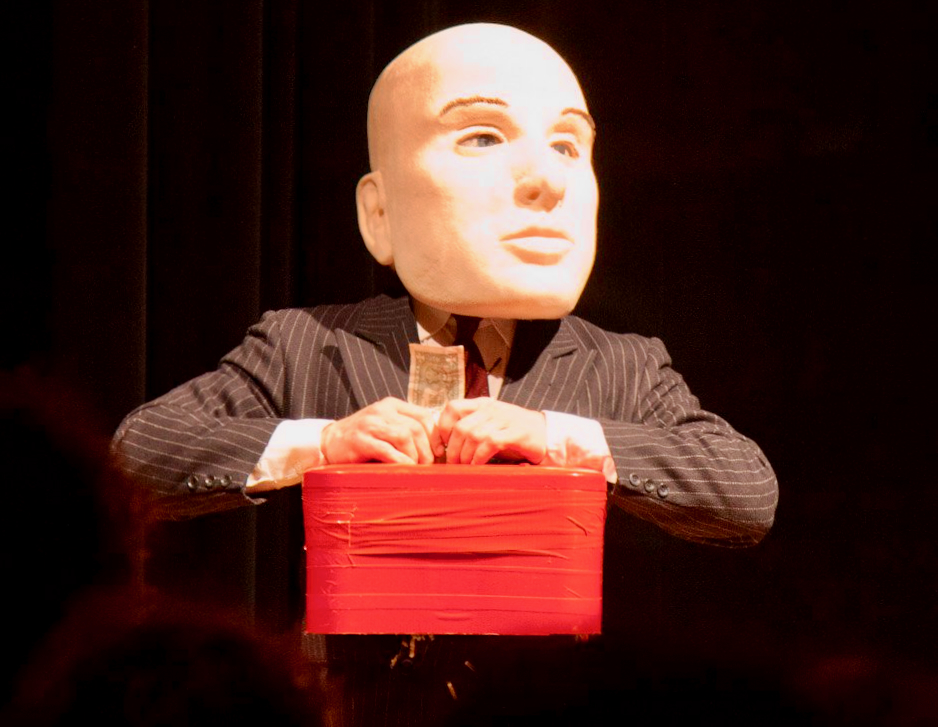 a man in a blue pinstripe business suit wears a giant bald human mascot head. attched to his chest is a red paper shredder and he is putting into it a dollar bill. 