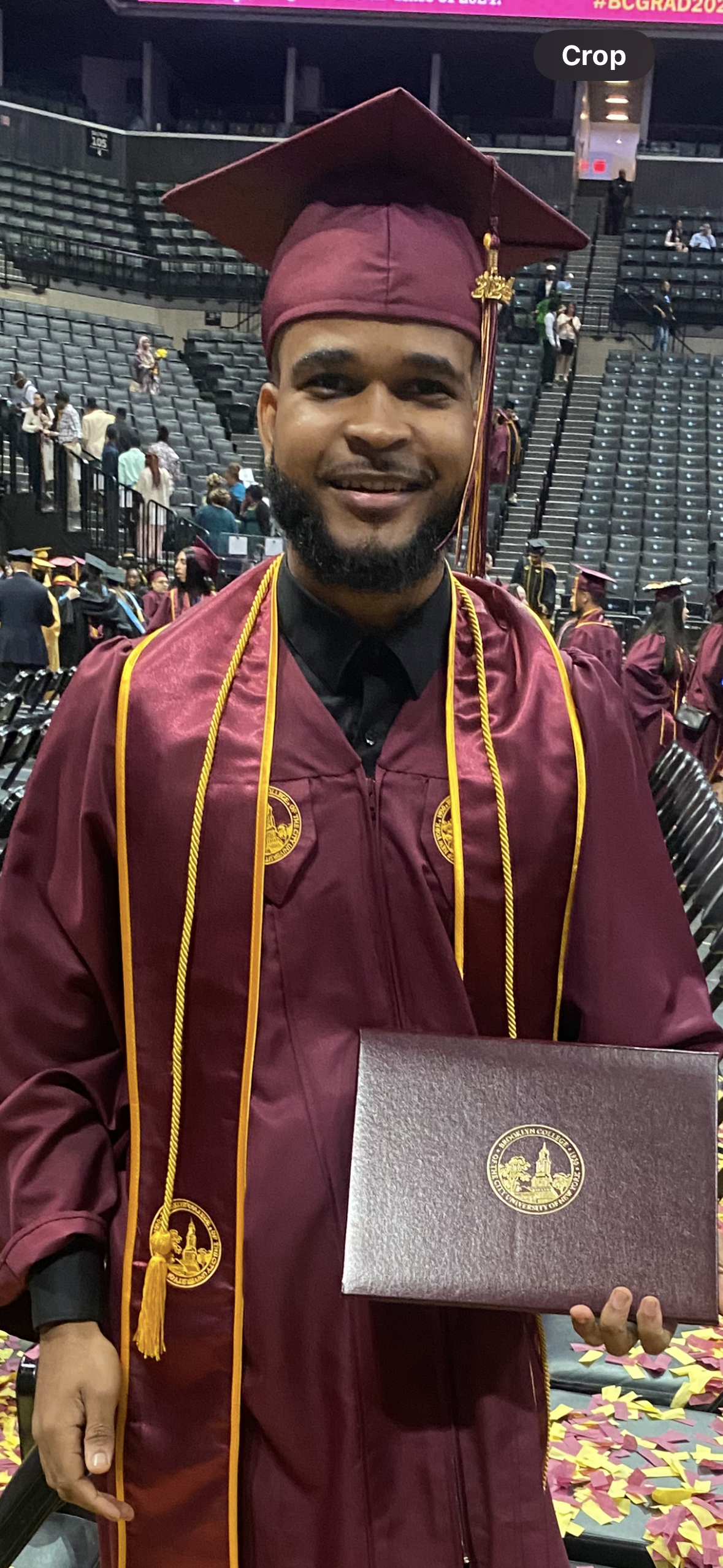 Clauny Cassagnol stands proudly in his cap and gown, holding his diploma with a beaming smile. He is surrounded by family members celebrating his graduation, showcasing their joy inside Barclays Center Stadium