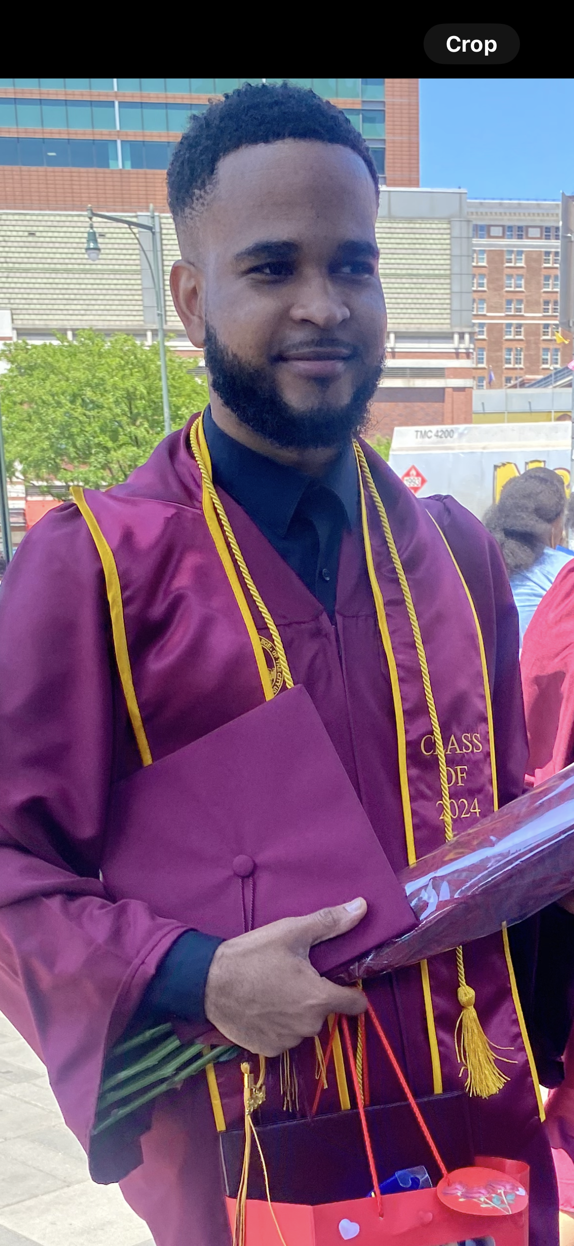 Clauny Cassagnol poses happily in his graduation gown and cap, clutching his diploma. The background features colorful balloons and proud family members, capturing the celebratory atmosphere of his academic achievement.