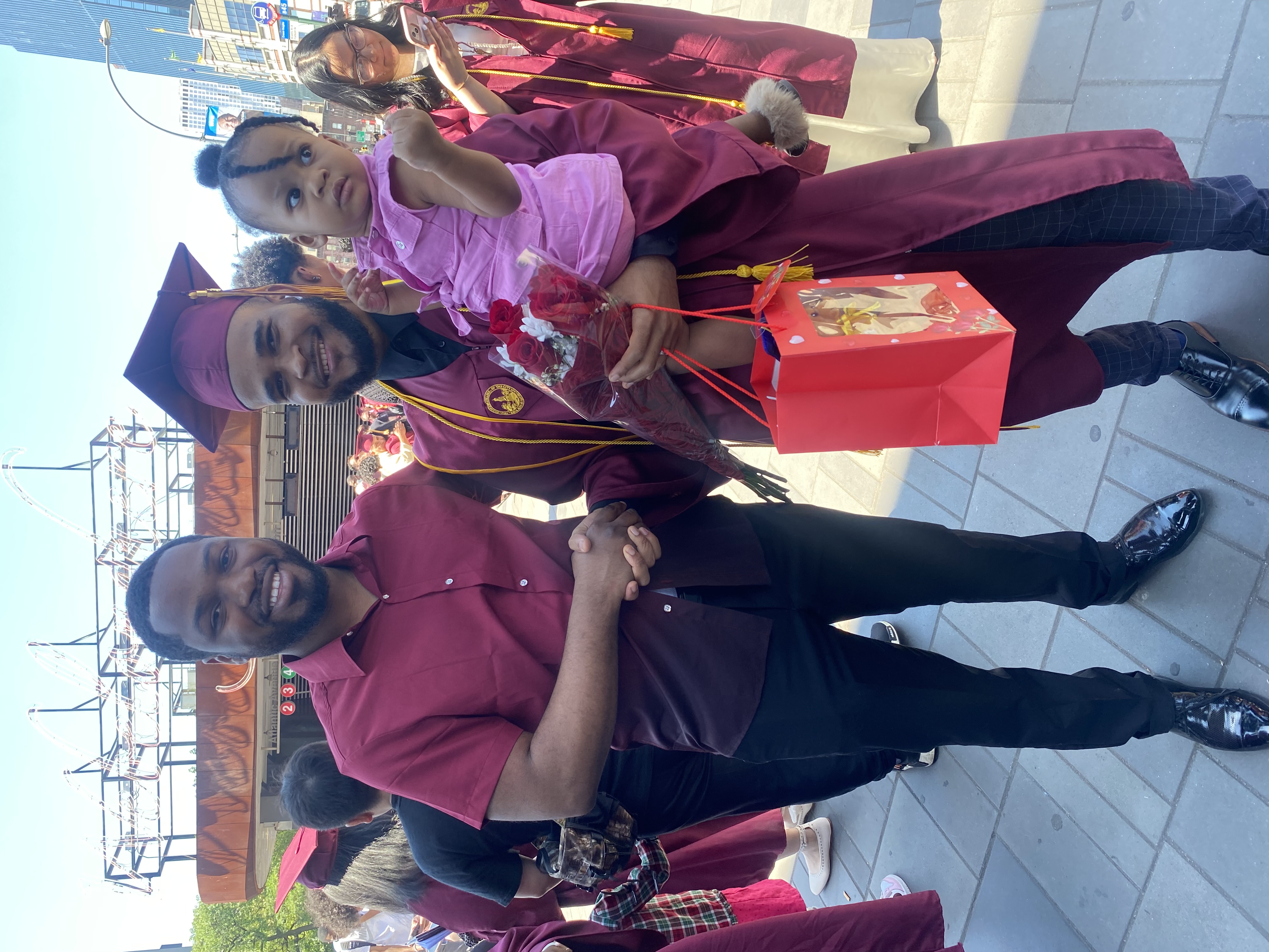 Clauny Cassagnol poses happily in his graduation gown and cap, holding his Dauighter and standing beside his friend