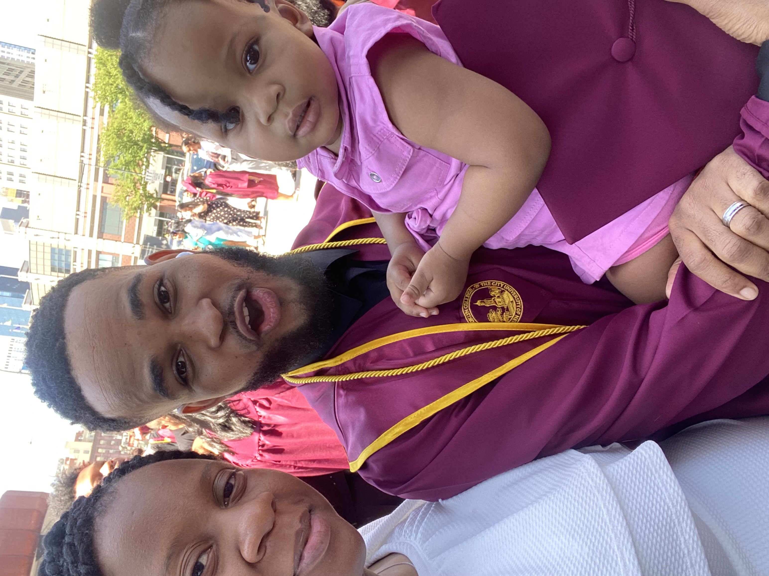 Clauny Cassagnol poses happily in his graduation gown and cap, holding his Dauighter and standing beside his Wife. 