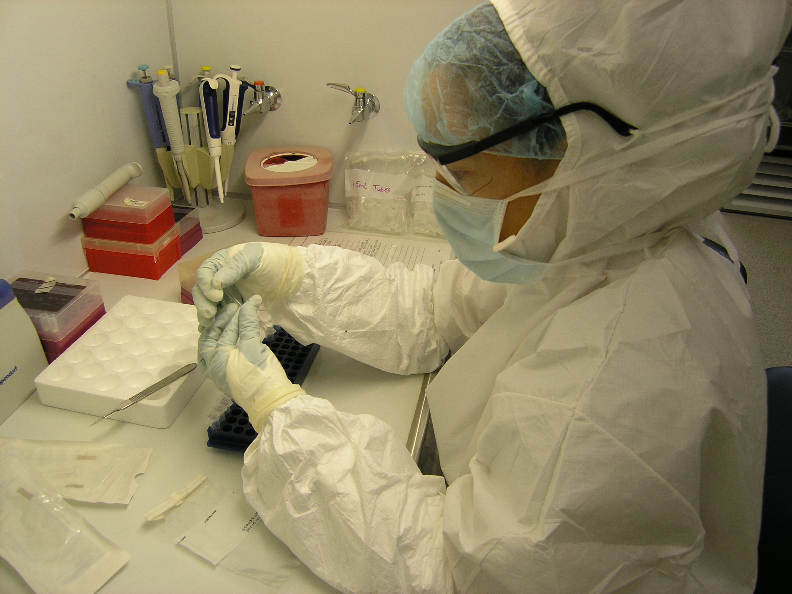Scientist in protective lab gear handling a sample tube at a biosafety workstation with pipettes and lab equipment.