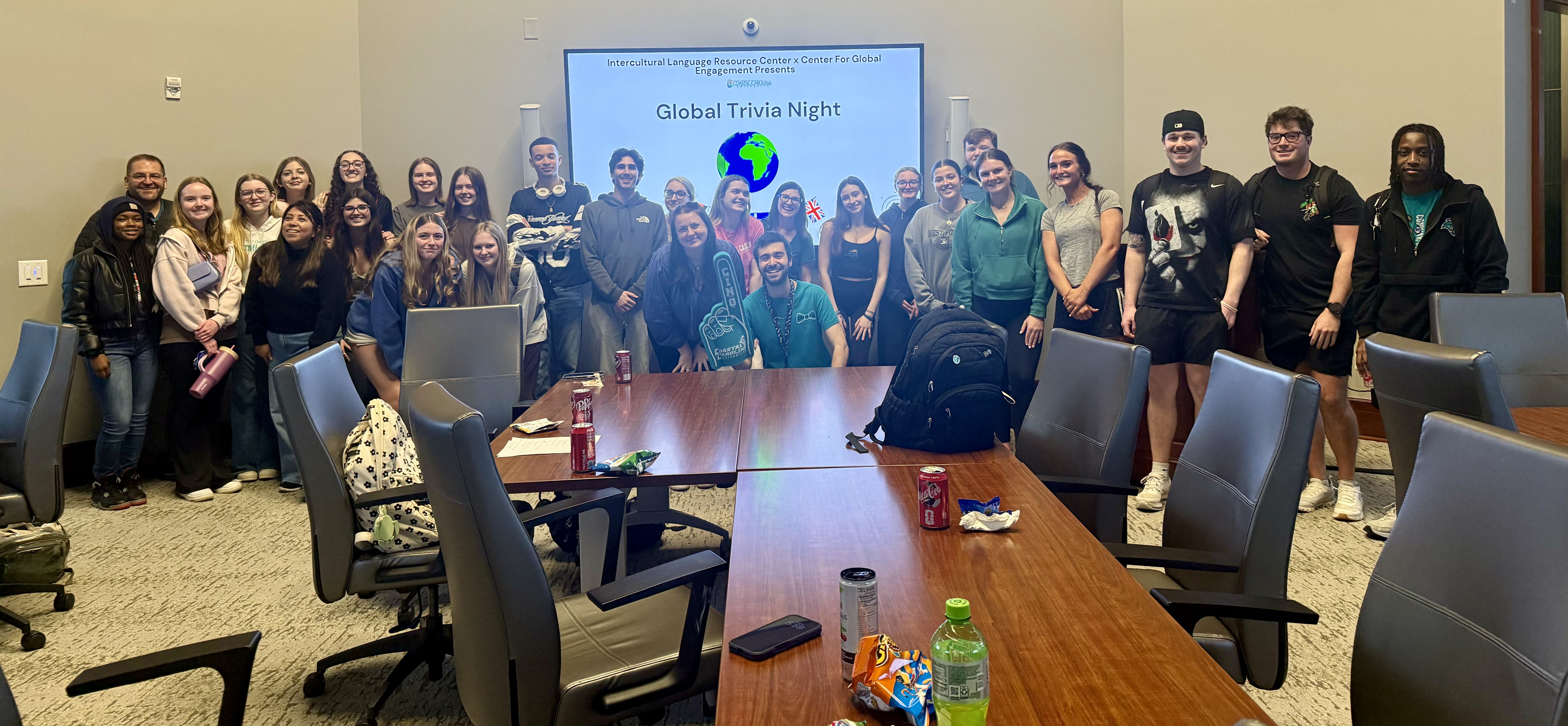 Participants stand around tables during a campus Global Trivia Night event, with a presentation displayed on a screen and students gathered for an intercultural activity.